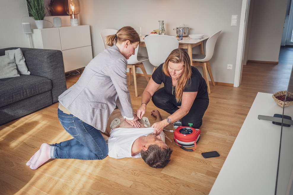 Zwei Frauen helfen einem kranken Mann, der am Boden liegt mit Herzmassage.